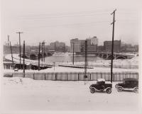 2 cars on snowy street with bridge and buildings in back